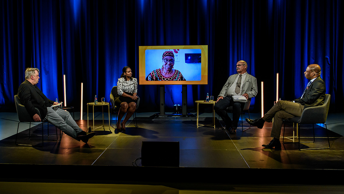The photo shows the panel with the moderator on the left and the three participants in the TV studio: Nessma Adil Yousif, Prof. Dr. Andreas Wrobel-Leipold and Dr. Gabin Ananou. The fourth participant, Dr Hedwig Ngwa from Cameroon, can be seen on a monitor in the middle behind the podium.