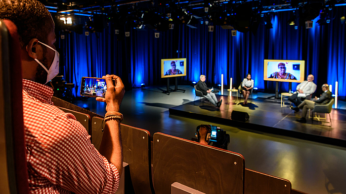 The photo shows a young man with dark skin looking over his shoulder at the „Dialog Kontrovers Extra“. He is filming from the audience stand with his smartphone into the TV studio, where four people on the podium and one person on a screen in the background can be seen. The focus is on the phone. 