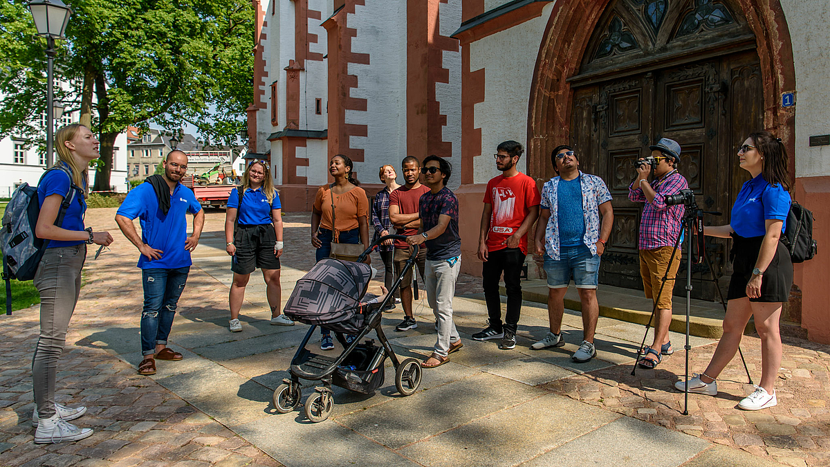 The photo shows a group of eleven young people in front of the side portal of the Protestant Town Church in Mittweida. Some are looking upwards. Four of them are wearing blue university shirts. They are the guides of the international group of students on their sightseeing tour of Mittweida.